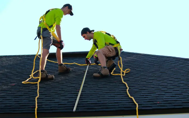 Two roofers in safety harnesses working on a dark shingle roof