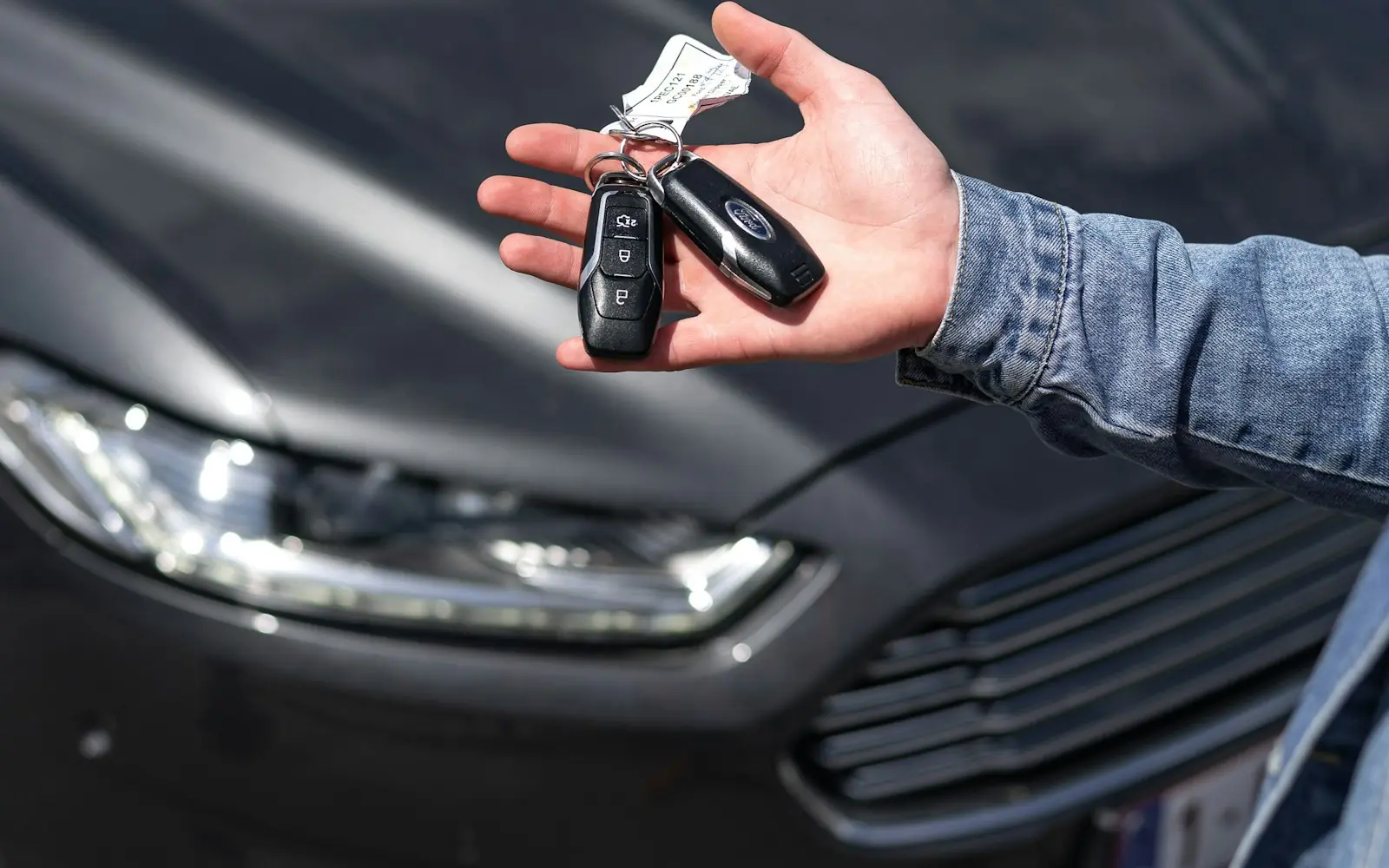 A hand holding a car key fob in front of a dark sedan
